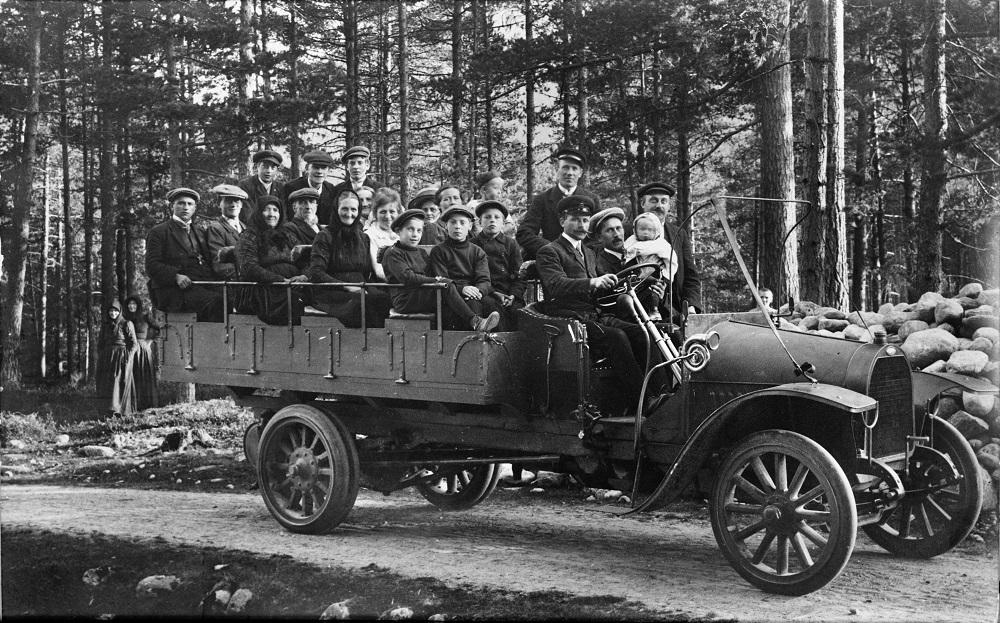 Dette var den første bilen A/S Fjordenes Automobilselskap kjøpte i 1913. Passasjerane er på veg til Breim, der dei skulle ta motorbåten inn til Førde. 21 personar sit på lasteplanet. Dei første bilane var små lastebilar. Når dei vart nytta til persontransport, vart det sett benkar eller stolar på lasteplanet. Olai Farsund, frå Førde, var sjåfør. Han var ein av dei dei første sjåførane i selskapet. 

 