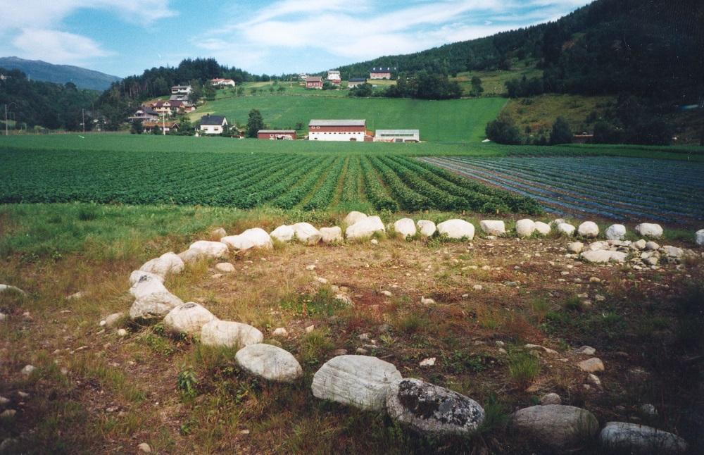 Biletet er teke på Vereidesletta ved den nye riksvegen. I framgrunnen er den såkalla Dronninggrava ramma inn med steinar. I bakgrunnen ser vi Bakketun-gardsbruket.
