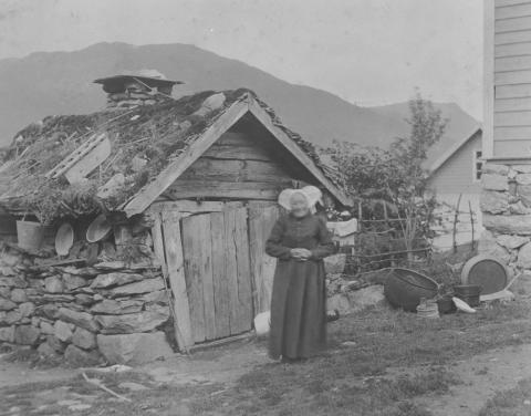 Frå tunet på plassen Munken. Huset til høgre er det noverande bustadhuset, bygt 1892/1893 og påbygt i 1937. Eldhuset t.v. vart rive i samband med påbygginga. Personen i biletet er truleg Brita Andersdotter (1827-1909). Huset i bakgrunnen er det gamle meieriet som vart sett opp kring 1898.
